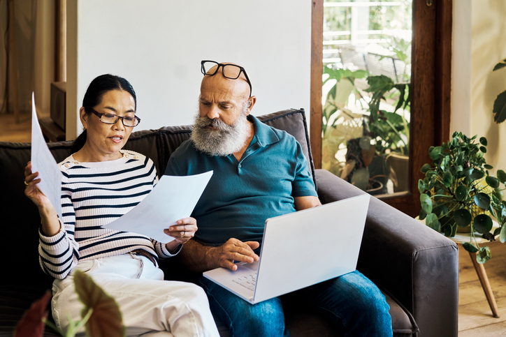 Man and women sitting on a couch. Man has a laptop. Woman is looking at papers.